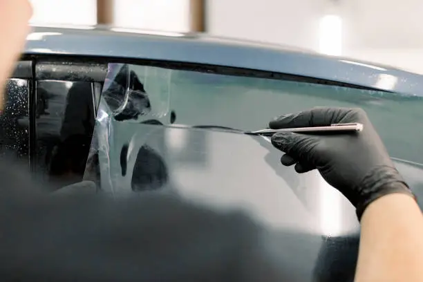 Photo of Cropped image of hands of worker in garage tinting a car window with tinted foil or film, holding special blade or knife to cut the film. Car detailing workshop, tinting windows