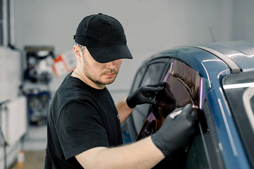 Imagen de primer plano de un trabajador mecánico de coches guapo, con uniforme negro, adjuntando papel de tintado a la ventana del coche en la estación de servicio especializada photo