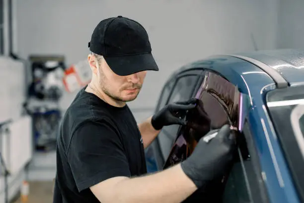 Photo of Closeup image of a handsome car mechanic worker, wearing black uniform, attaching tinting foil to car window in specialized service station