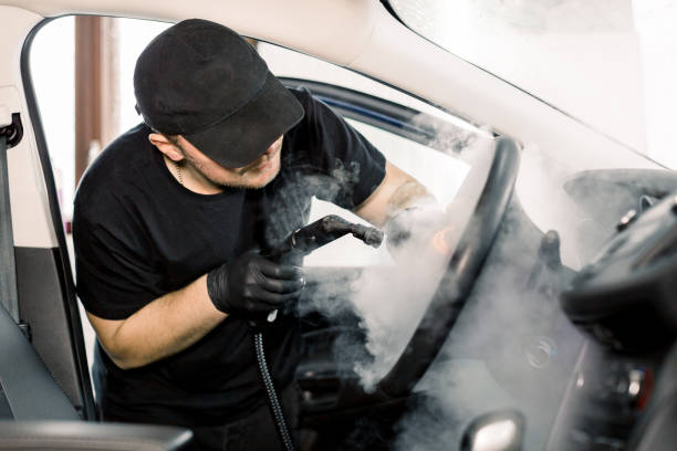 auto-detaillierung, dampfreinigungskonzept. hübscher mann in schwarzem t-shirt und mütze, arbeiter von autowaschzentrum, reinigung auto innenraum mit heißen dampfreiniger. auto-detaillierungskonzept - blank shirt stock-fotos und bilder