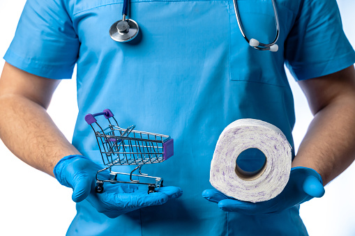 Young male doctor with stethoscope and protective face mask showing shopping cart with toilet paper on white background, close-up. The scene is situated in controlled photography studio environment in front of white background. Picture is taken with Sony A7III camera.