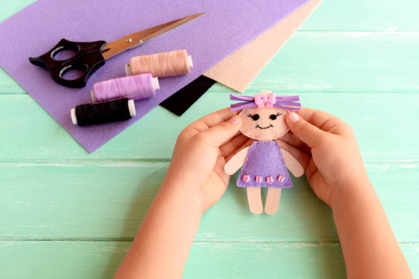 istock A child holds a small doll in his hands. Doll is sewn from felt. Scissors, thread and felt set on a wooden table. Cute fabric art and craft idea for kids 1220583739