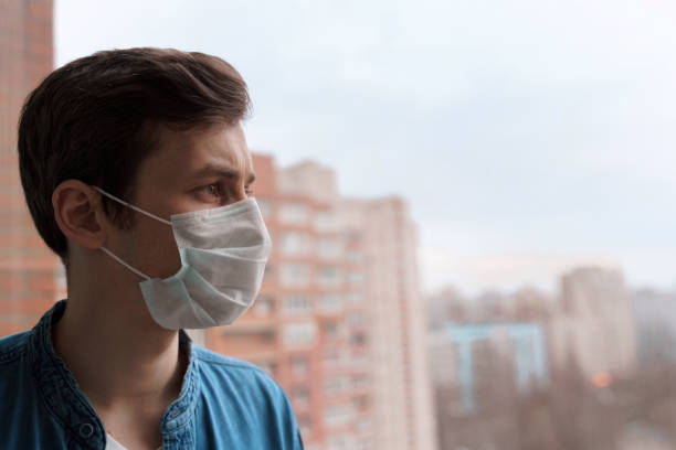 istock Depressed man in protective mask looking out the window 1220588202