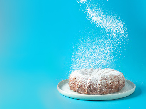 Woman's hand sprinkling icing sugar over fresh home made bundt cake. Powder sugar falls on fresh perfect bunt cake over blue background. Copy space for text. Ideas and recipes for breakfast or dessert
