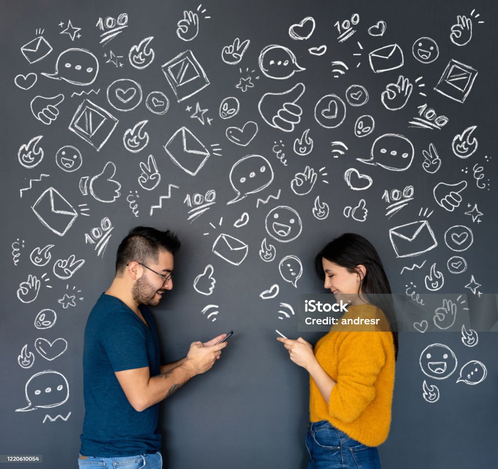 Happy young couple texting on their cell phones Happy young couple texting on their cell phones with icons on a blackboard behind them Emoticon Stock Photo