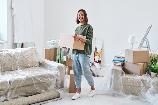 Cheerful young woman in jeans and sweatshirt carrying packed box while moving along new flat or house in front of camera