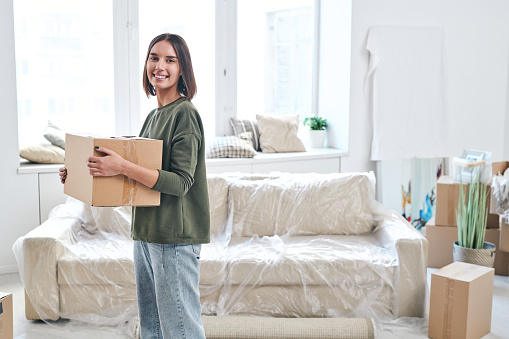 Happy young woman with packed box looking at you with toothy smile while standing on background of couch and window