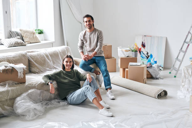 istock Cheerful young man and woman in jeans and pullovers sitting by couch 1220616813