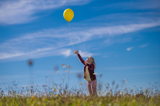 Little girl releasing yellow balloon against blue sky while standing in field.