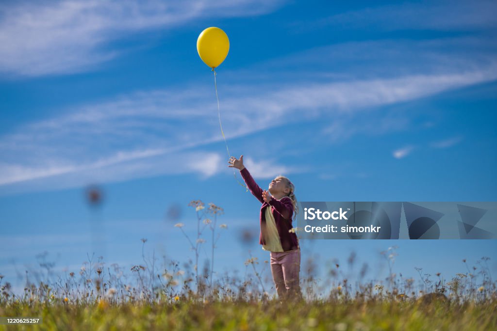 Girl releasing balloon against blue sky Little girl releasing yellow balloon against blue sky while standing in field. Balloon Stock Photo