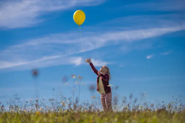 Little girl releasing yellow balloon against blue sky while standing in field.