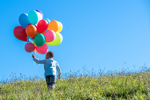 Rear view of boy holding bunch of colourful balloons against blue sky while walking in field.
