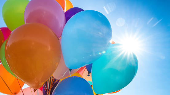 Low angle view of bunch of multicoloured balloons and sun flares against blue sky.