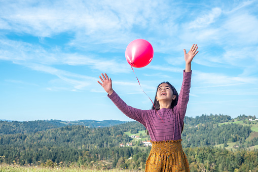 Girl releasing balloon against sky with lushy mountains in background.