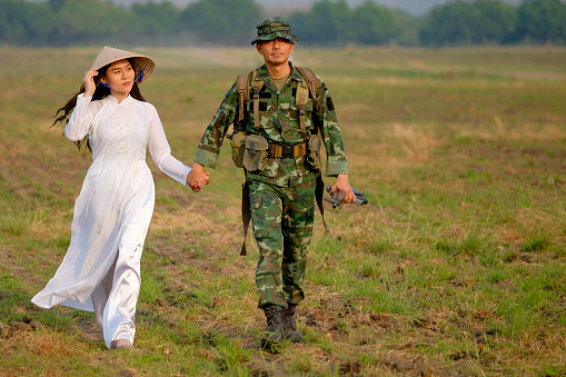 Soldier man holds hand of Vietnamese woman and walk in the field with evening light in concept of love during war.