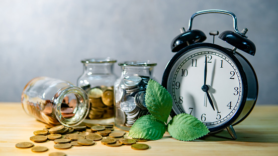 Clock with coins in currency glass jars and spilling on wooden table. Saving money for future retirement. Financial business growth. Time investment concept