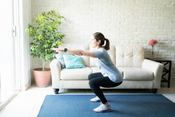 Active woman doing squats at home Full length view of an active woman in her 40s working out and doing some squats at home squatting position photos stock pictures, royalty-free photos & images