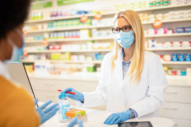 Pharmacist with protection mask giving instructions on how to use medicine to a client. Female healthcare worker explaining to a client with virus symptoms how to use a medicine in pharmacy house. chemist photos stock pictures, royalty-free photos & images
