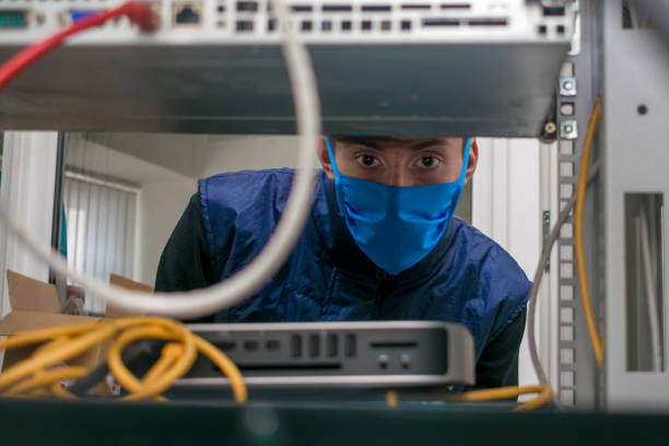 a technician in a medical mask works in a rack with computer equipment. the man serves the server room in the datacenter. portrait of a teenager in a data center. - network server computer tower rack imagens e fotografias de stock