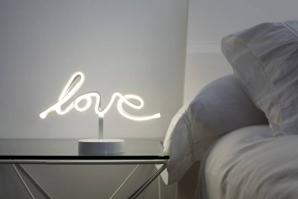 Love and Togetherness Through Objects: Love Lamp, Night Time Modern, clean style, white bedroom photographed in the evening. This couple's bedroom has a glass bedside table with a bright, love shaped, neon lamp. Cozy bed with big, comfortable pillows and white linen. neon bedroom stock pictures, royalty-free photos & images