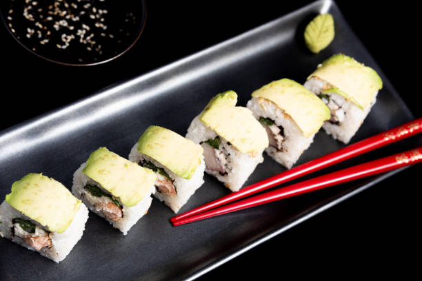 sushi roll with avocado wrap, grilled salmon, philadelphia cheese and green onions on black plate and red chopsticks. small pot with soy sauce. black background. - fine dining grilled spring onion healthy lifestyle imagens e fotografias de stock