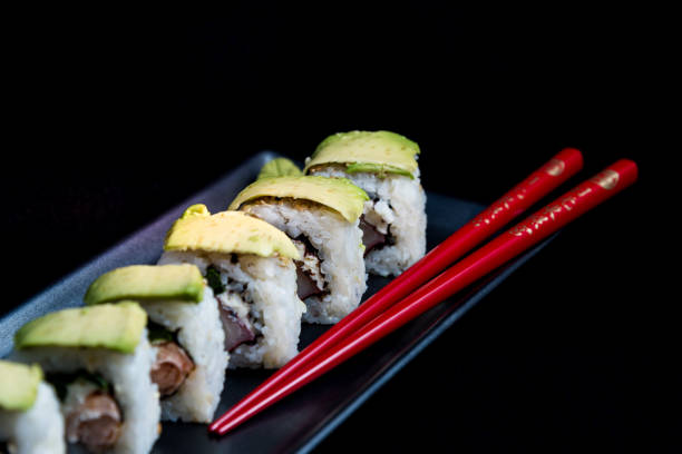 sushi roll with avocado wrap, grilled salmon, philadelphia cheese and green onions on black plate and red chopsticks. small pot with soy sauce. black background. - fine dining grilled spring onion healthy lifestyle imagens e fotografias de stock