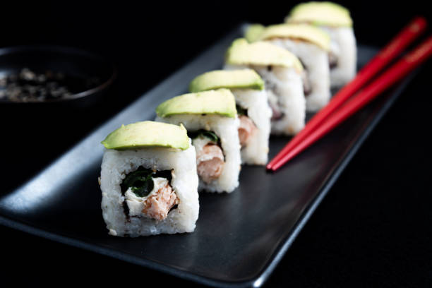 sushi roll with avocado wrap, grilled salmon, philadelphia cheese and green onions on black plate and red chopsticks. small pot with soy sauce. black background. - fine dining grilled spring onion healthy lifestyle imagens e fotografias de stock