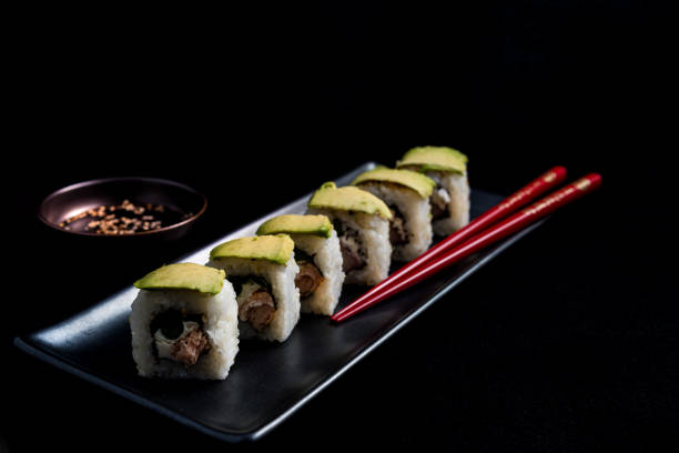 sushi roll with avocado wrap, grilled salmon, philadelphia cheese and green onions on black plate and red chopsticks. small pot with soy sauce. black background. - fine dining grilled spring onion healthy lifestyle imagens e fotografias de stock