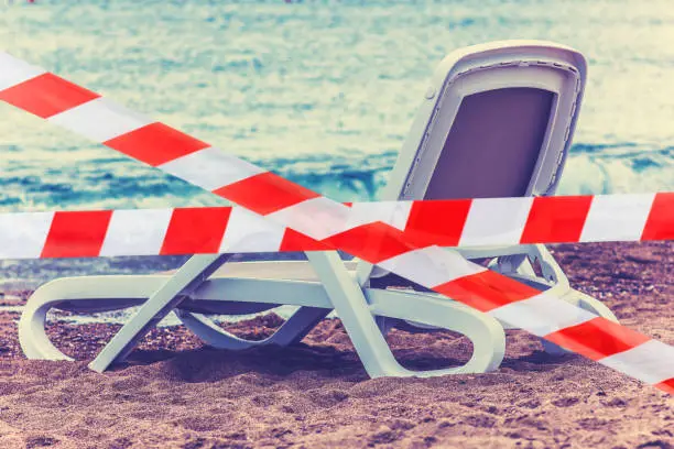 Photo of The beach lounger is blocked with a red and white ribbon, the concept of canceling holidays or canceling summer plans due to a pandemic and viral diseases.