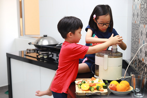 Cute Asian sibling making fruit juice using electric juicer in the kitchen at home