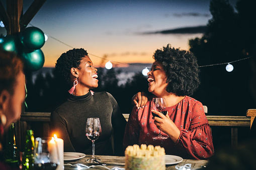 Female friends laughing while talking at table. Women are enjoying wine at birthday party. They are in illuminated balcony during night.