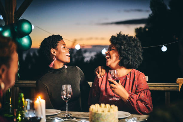 Cheerful female friends enjoying wine at birthday Female friends laughing while talking at table. Women are enjoying wine at birthday party. They are in illuminated balcony during night. public lighting stock pictures, royalty-free photos & images