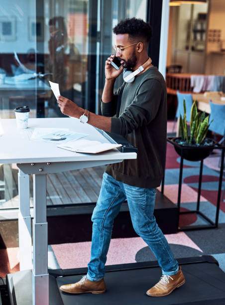 The multitasking pro is at it again Shot of a young businessman talking on a cellphone and going through paperwork while walking on a treadmill in an office standing desk photos stock pictures, royalty-free photos & images