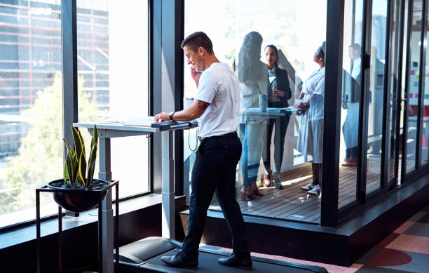 Walk and work your way to success Shot of a young businessman talking on a cellphone and going through paperwork while walking on a treadmill in an office standing desk photos stock pictures, royalty-free photos & images
