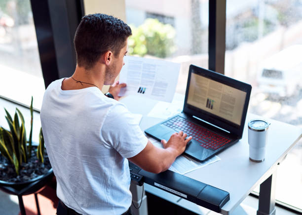 Sit less and walk more Shot of a young businessman going through paperwork and working on a laptop while walking on a treadmill in an office standing desk photos stock pictures, royalty-free photos & images