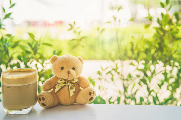 istock Cute brown teddy bear sitting on white table side a cup of coffee next a glass window seeing  nature outside background. 1220706485