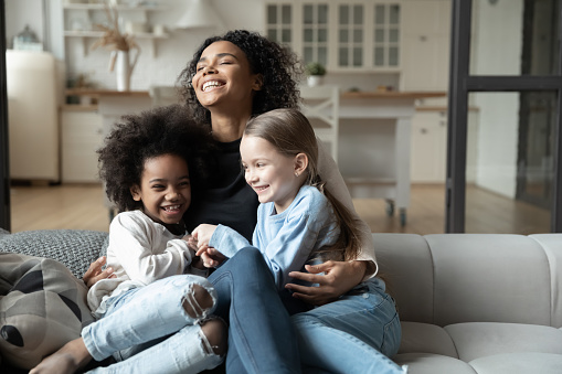 Overjoyed young African American mother play with little multiracial daughters at home, happy biracial mom sit on couch tickle have fun with smiling small multiethnic girls kids on weekend together