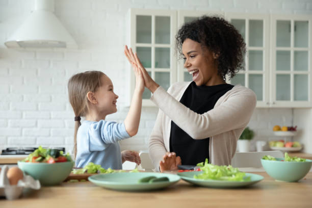 Happy multiethnic mom and little daughter have fun cooking Overjoyed young African American mom and little Caucasian daughter give high five cooking at home together, smiling biracial mother and small girl child have fun preparing healthy food in kitchen nanny stock pictures, royalty-free photos & images