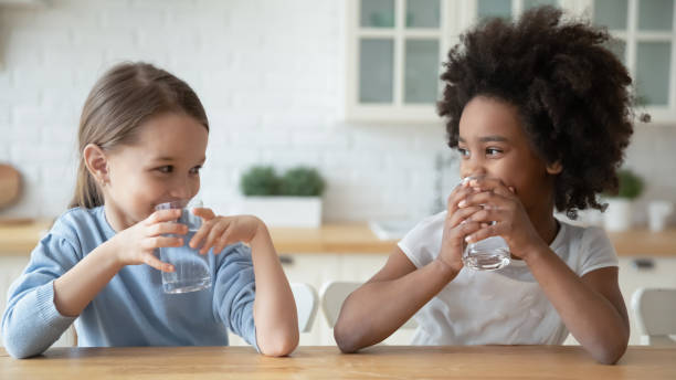pequeñas chicas multiétnicas beben agua mineral en casa - agua potable fotos fotografías e imágenes de stock