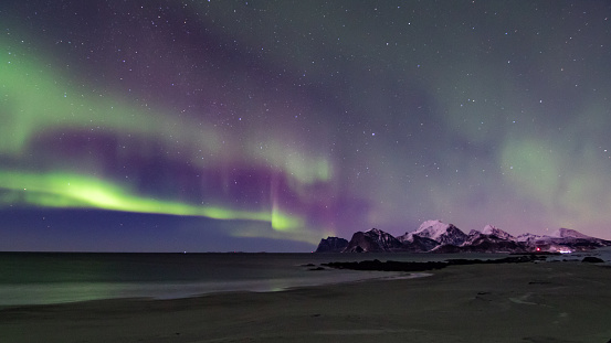 In the foreground, the beach reflects the glow in the sky. In the background, the snow-covered mountains.