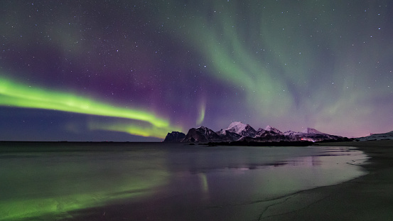 In the foreground, the beach reflects the glow in the sky. In the background, the snow-covered mountains.