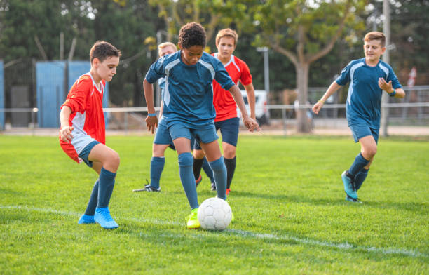 Red and Blue Jersey Boy Footballers Competing on Field Action portrait of 13 year old blue jersey footballer controlling ball against red jersey teammate during practice match. soccer team stock pictures, royalty-free photos & images