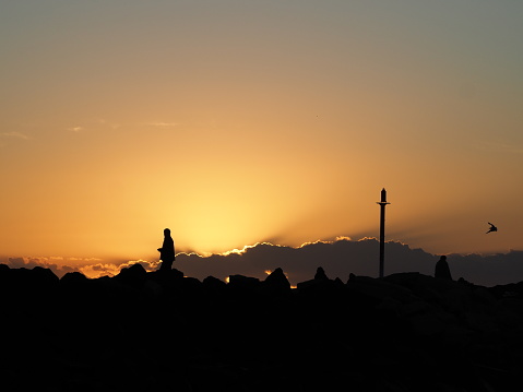 Unidentifiable human figures in silhouette, standing on the sea rock wall at Brunswick Heads, NSW contrast sharply with the golden sunrise sky behind them during the time of the covid 19 pandemic.