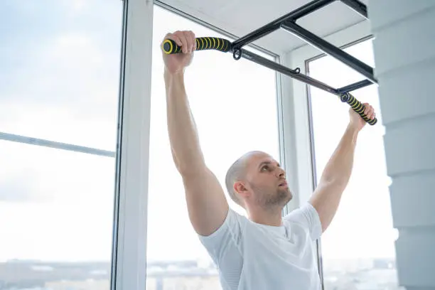 Man goes in for sports doing pull-up exercises on horizontal bar at his home.