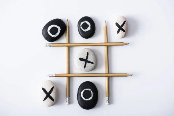 top view of tic tac toe game with grid made of pencils, and pebbles marked with crosses and naughts on white surface