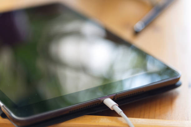 Close-up digital tablet charging on wooden work table. Black pen blurred in the background. Leaf reflections on the tablet screen. Horizontal.