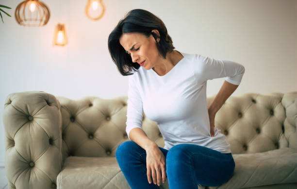 Axial pain. Close-up photo of a hurting woman, who is sitting on a couch and holding her lower back with her left hand. stock photo