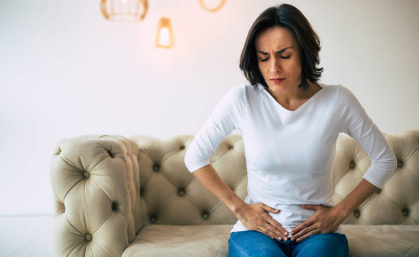 problemas femeninos. una mujer adulta está sentada en un sofá en casa y tocándose la parte inferior del estómago mientras sufre de calambres. - holding belly fotografías e imágenes de stock