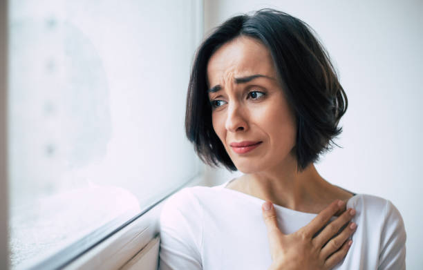 enfermedad grave. foto de primer plano de una joven que está mirando por la ventana y llorando, mientras sostiene su mano sobre su pecho. - pessimist women one person caucasian fotografías e imágenes de stock