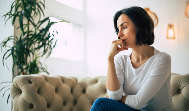 pensando en mis pensamientos. foto de primer plano de una mujer seria, que está sentada en un sofá en su apartamento moderno y pensando, mientras se toca la barbilla con la mano derecha. - pessimist women one person caucasian fotografías e imágenes de stock